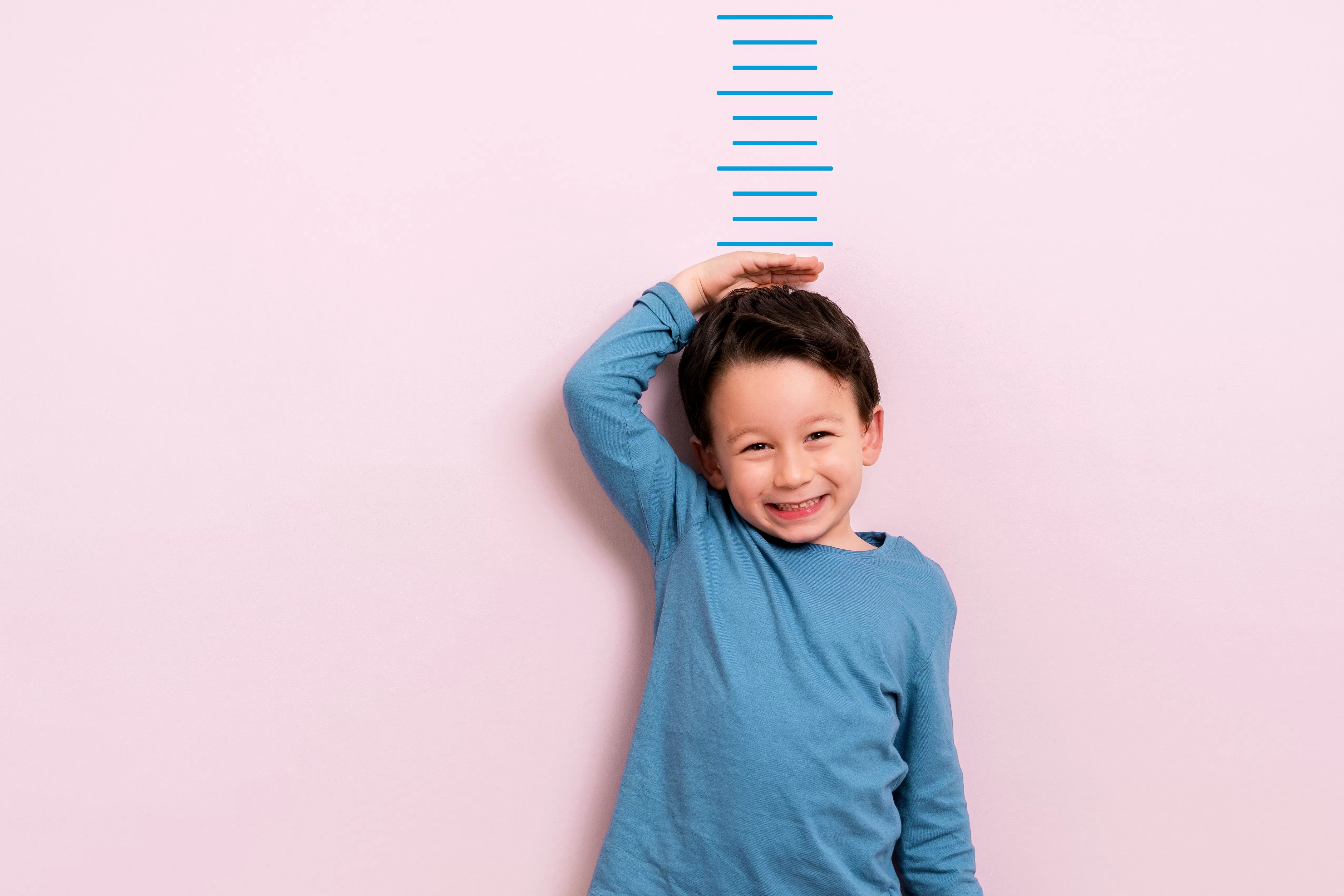 Criança de camisa azul encostado em uma parede rosa com desenho de uma régua medindo seu tamanho 